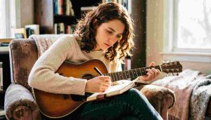 A singer-songwriter playing acoustic guitar in a softly lit room while writing lyrics in a notebook